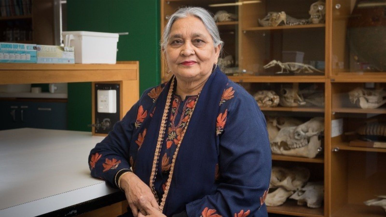 Indian woman wearing a navy cloak with orange flowers, with skulls of animals in a cabinet behind