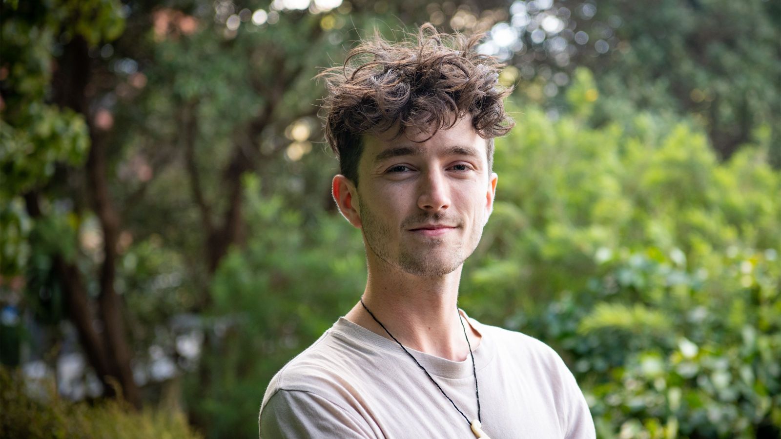 man with curly hair looking at camera, green leafy background