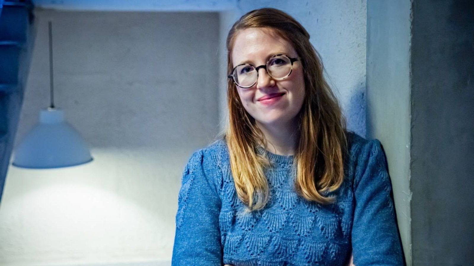 Girl with long red hair and blue jumper in concrete gallery with blue lamp