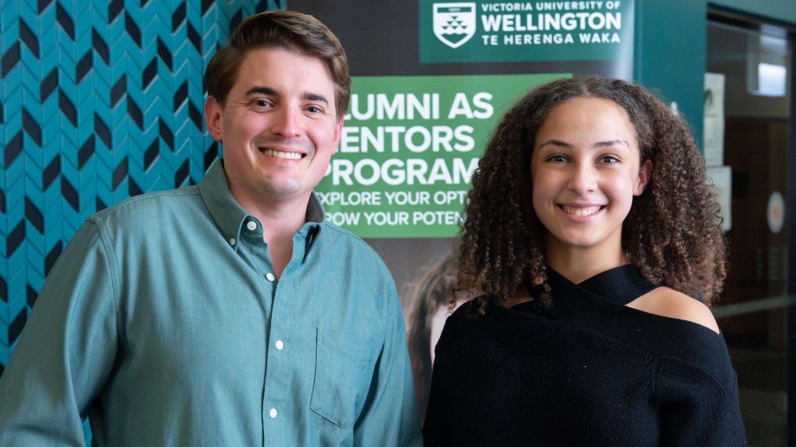 two people looking at camera one with short brown hair, one with long curly hair against alumni as mentors banner