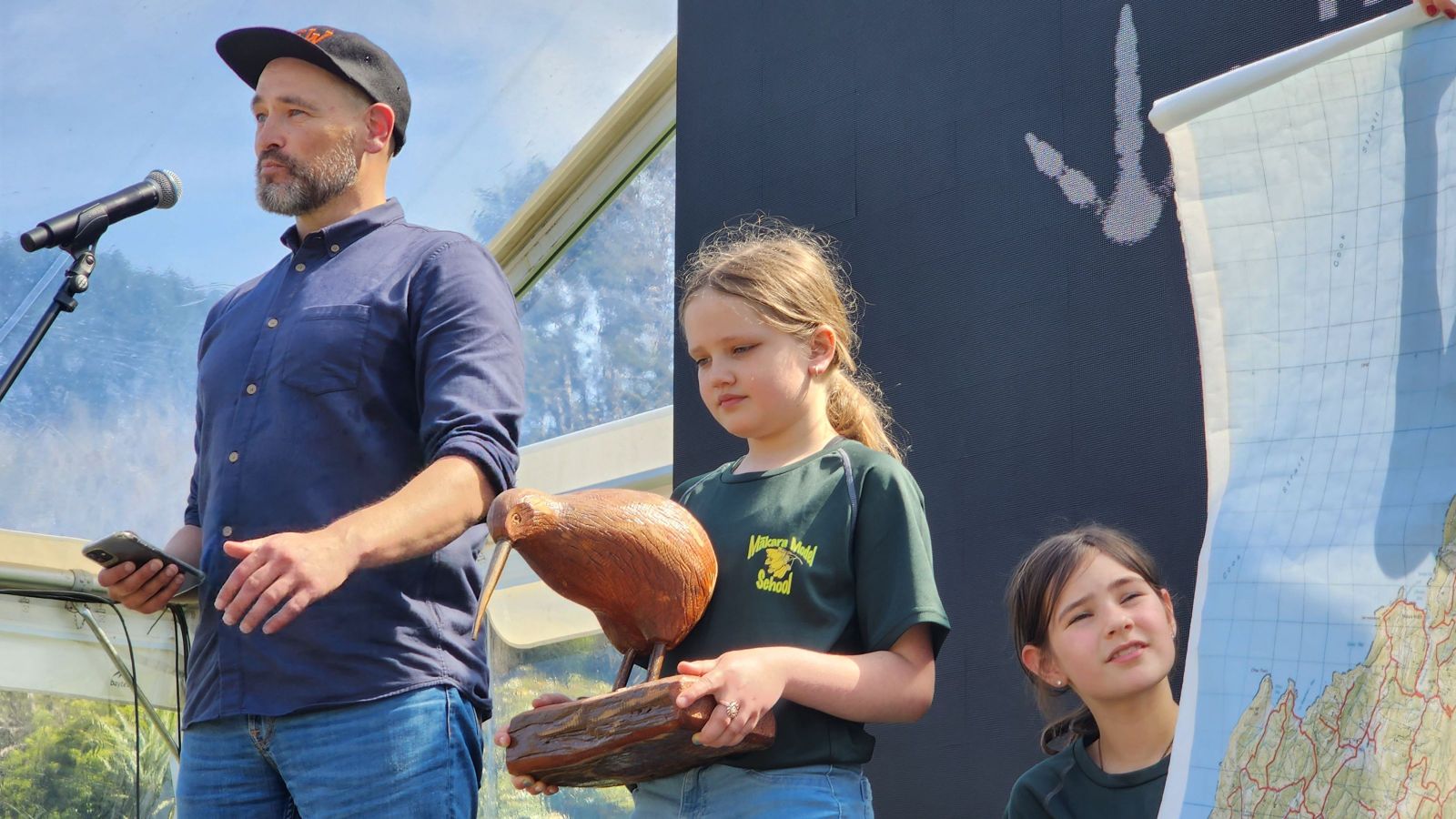 man at microphone with girl beside him holding a wooden kiwi