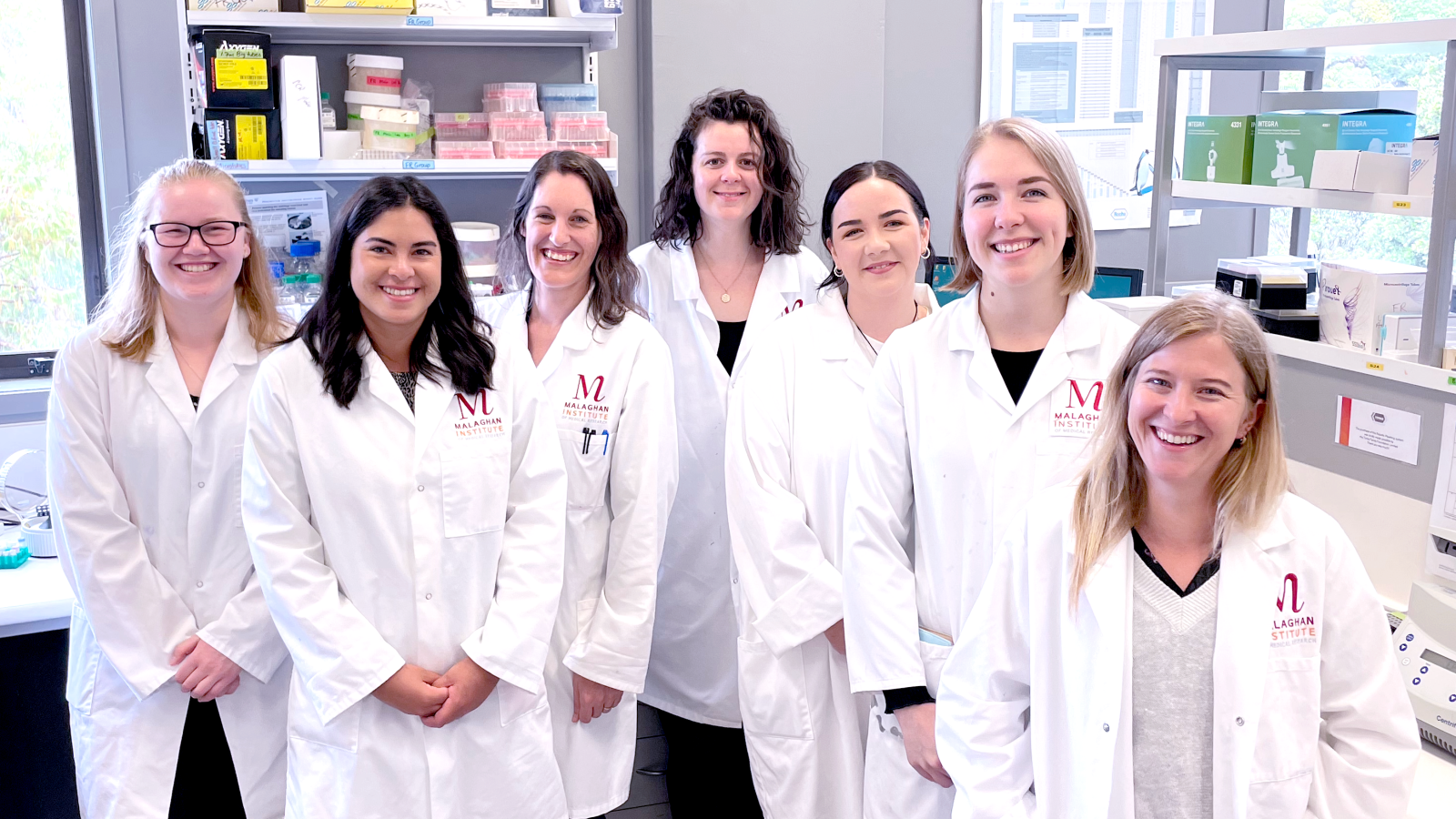 A group of women wearing labcoats stand smiling at the camera.