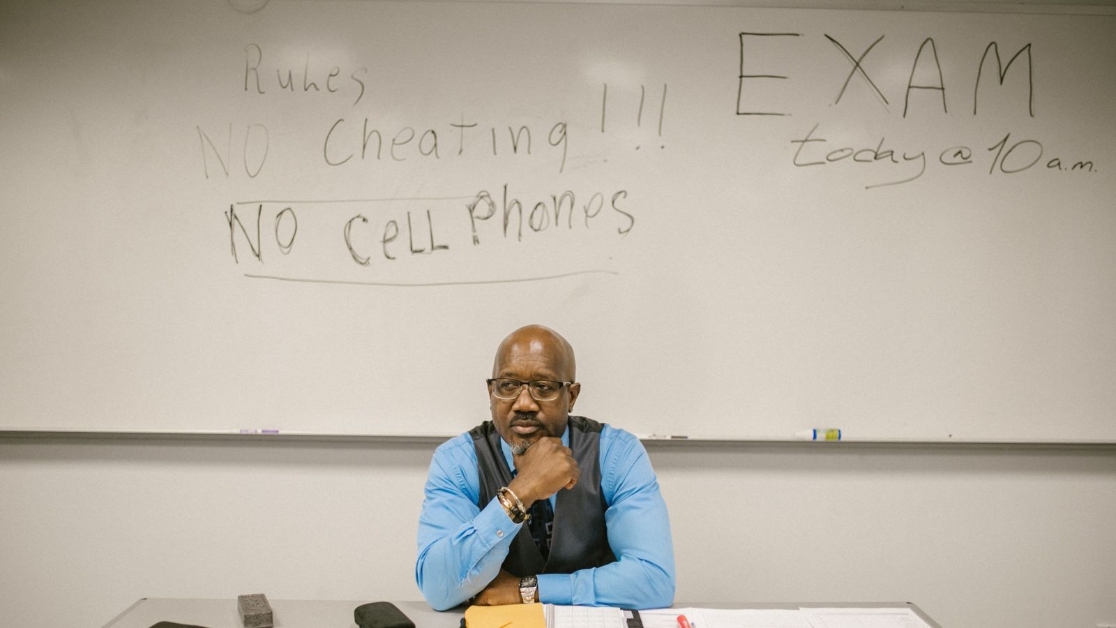 Teacher sitting at desk with whiteboard behind, reading 