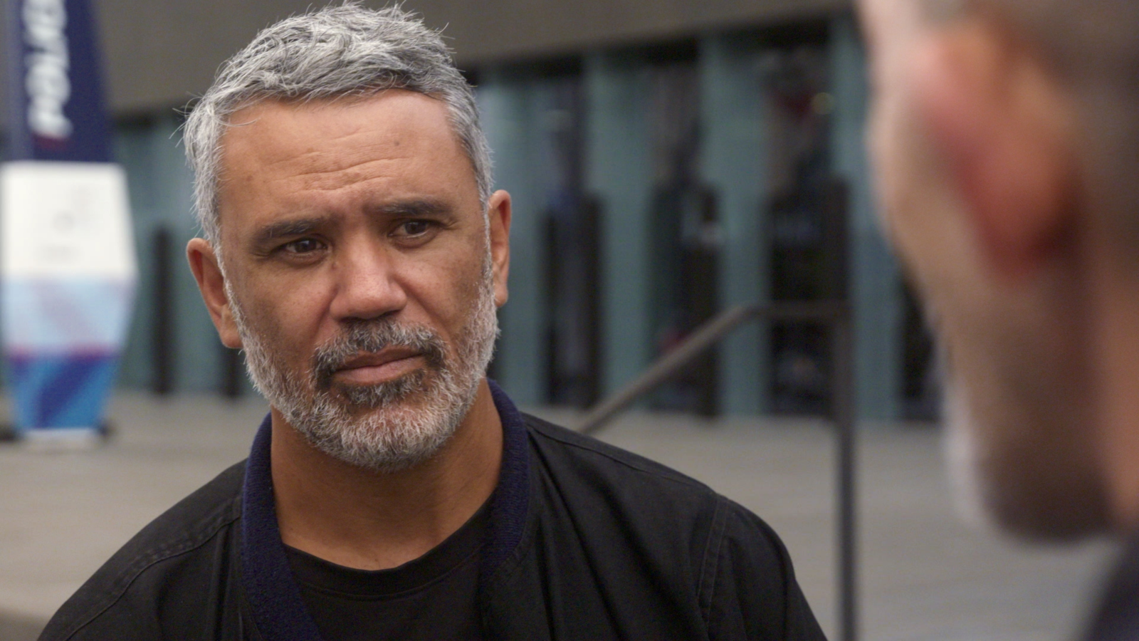 A Māori man stands staring into the camera.