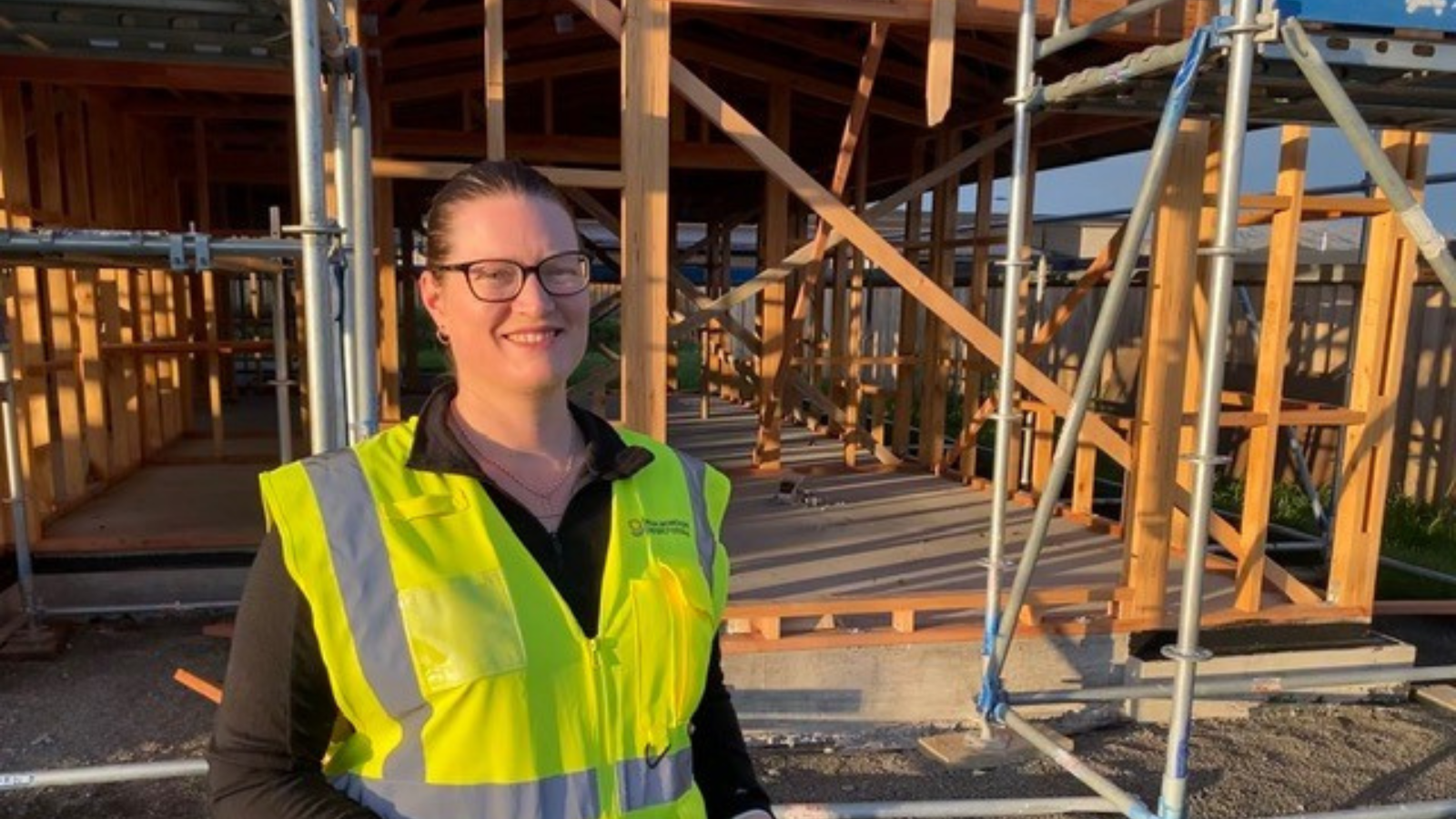 An adult woman holds a clipboard in front of a house under construction.