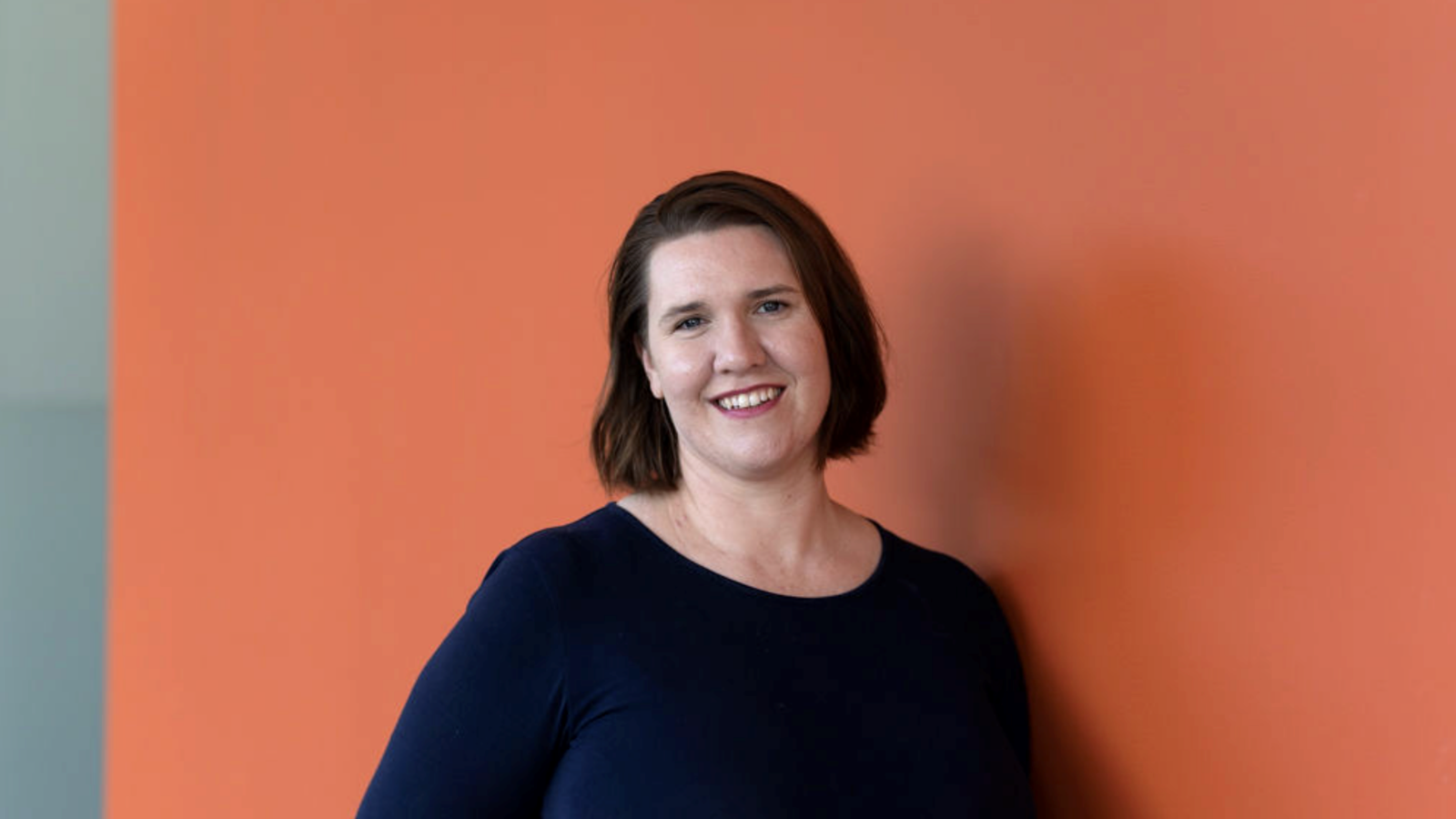 A young woman stands in front of an orange wall smiling at the camera.