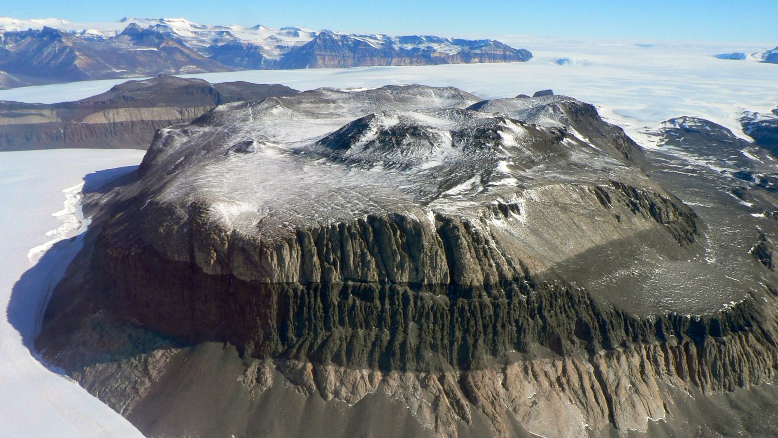 A glacier in antarctica. There is blue sky in the background and a large mountain with a dark surface and snow on the top.