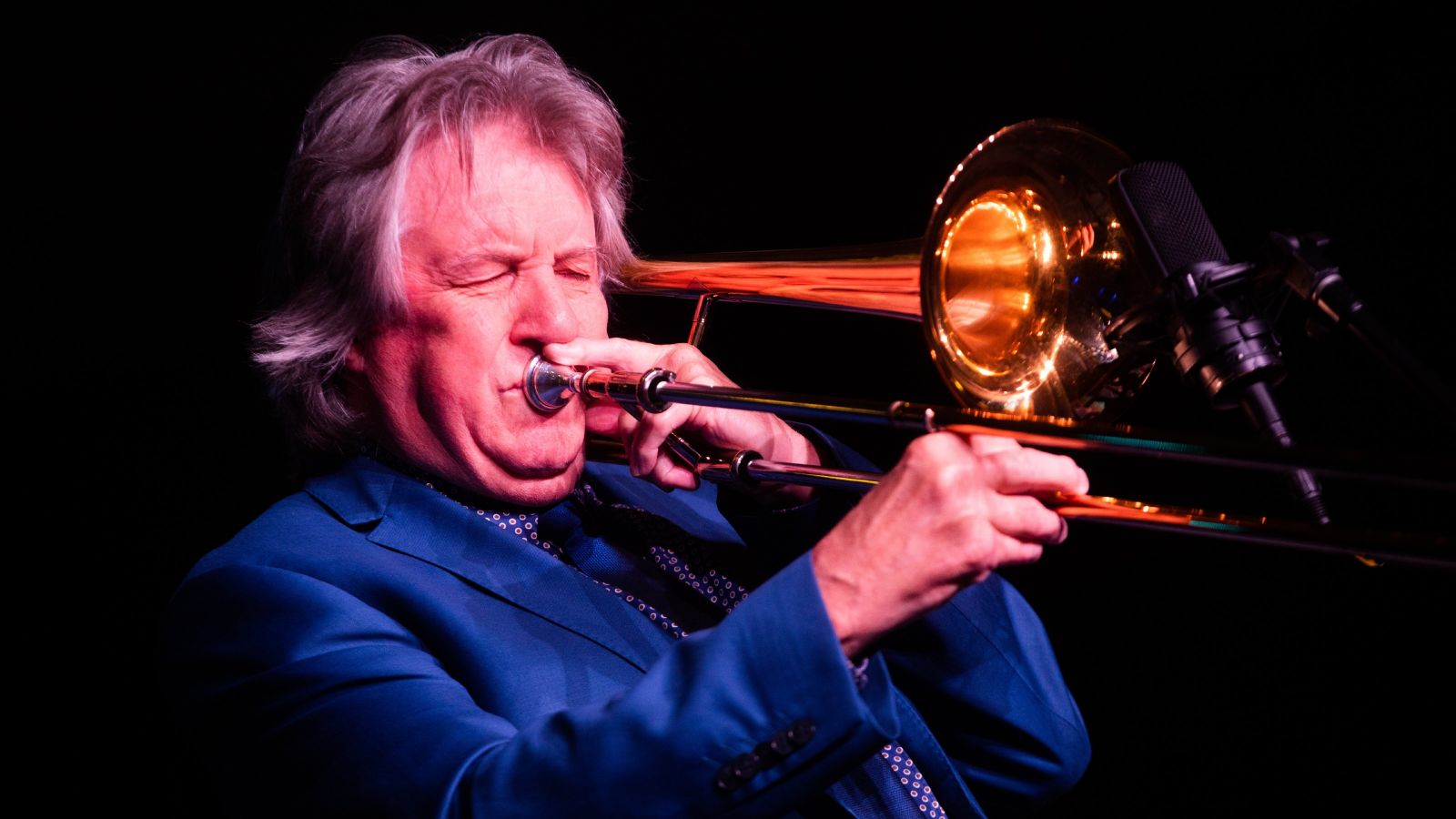 Rodger Fox is wearing a blue suit and playing a trumpet.