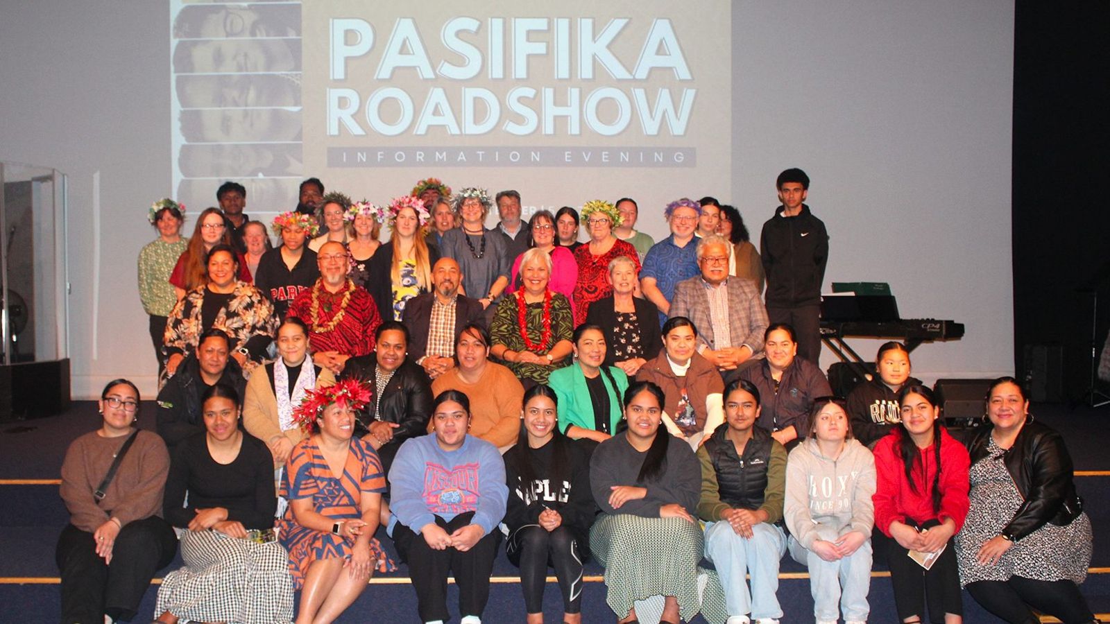 A large group of people in a room sitting in rows with a sign for the Pasifika Roadshow behind them.