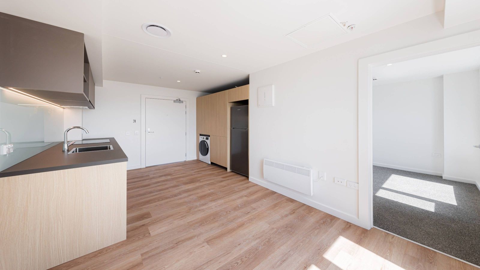 A one bedroom apartment, there is a kitchen on the left, and a washing machine and overlook of the dining area on the right. The walls are white, and there is wood flooring,
