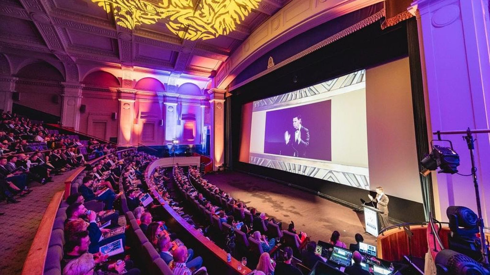 Alumnus Patrick Gower stands behind a lectern on stage as he hosts the evening to the large gathered crowd  