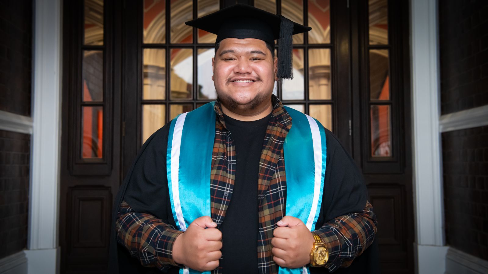 samoan man with plaid shirt and light blue scarf with white edging, standing proudly between two white poles