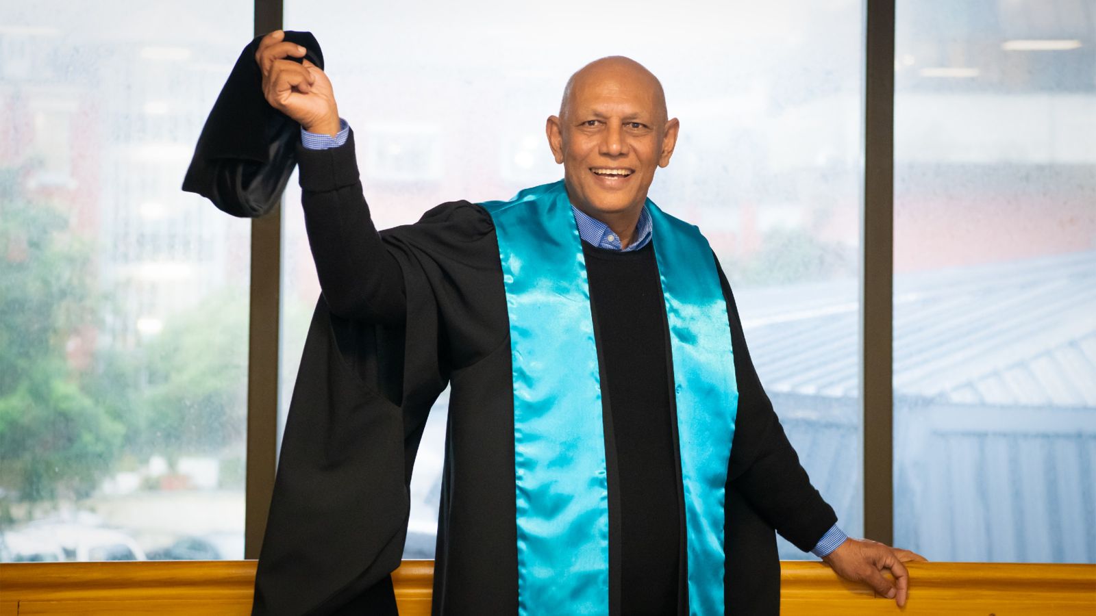 Dr Anoop Kumar stands in front of a window holding his grad cap up proudly. 