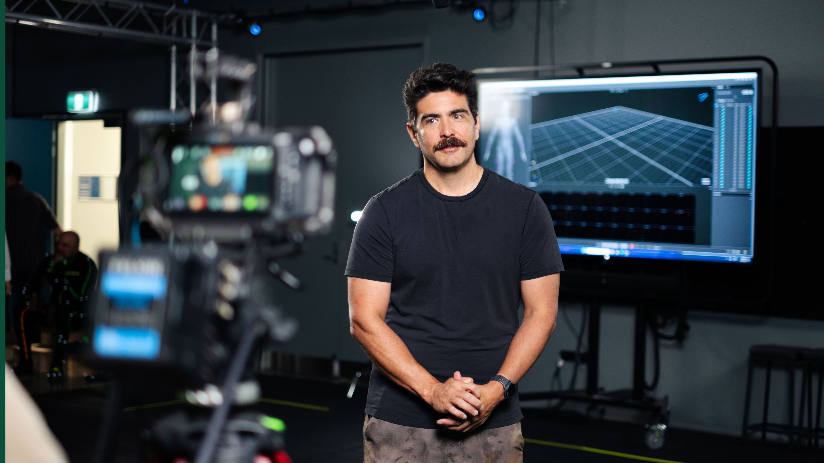 moustachioed man in black tshirt standing in front of graphic rendering on tv