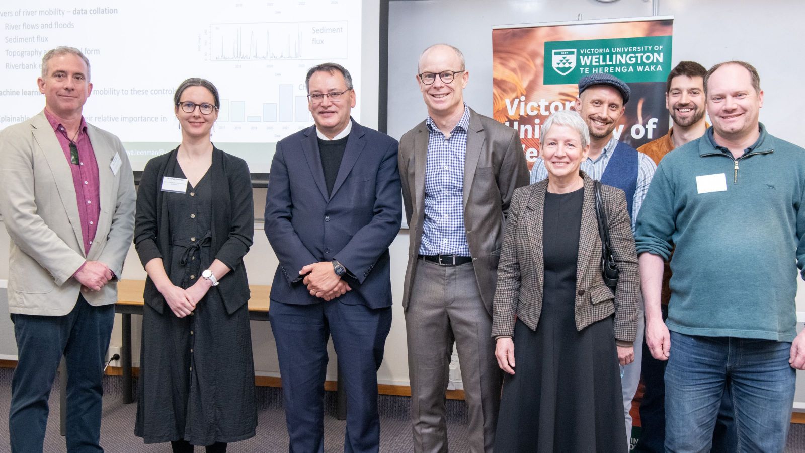 Eight people posing for a photo with a banner with Victoria University of Wellington in back