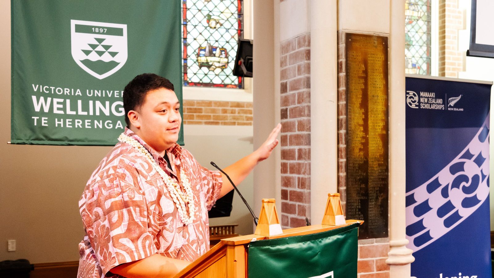 Man in pink and red patterned shirt wearing shell necklace gesturing behind a podium