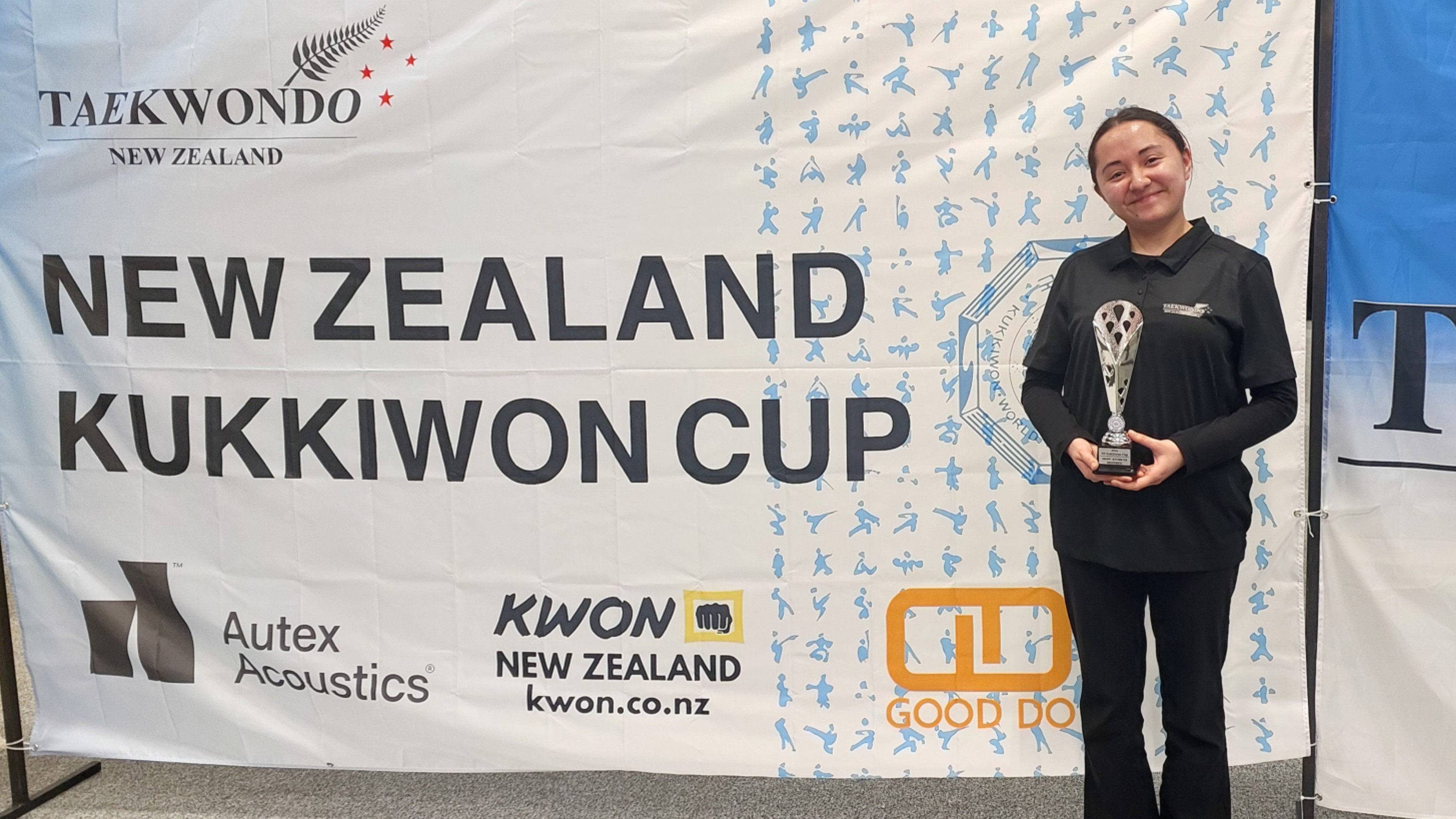 Young woman smiles as she holds a trophy in front of competition signage