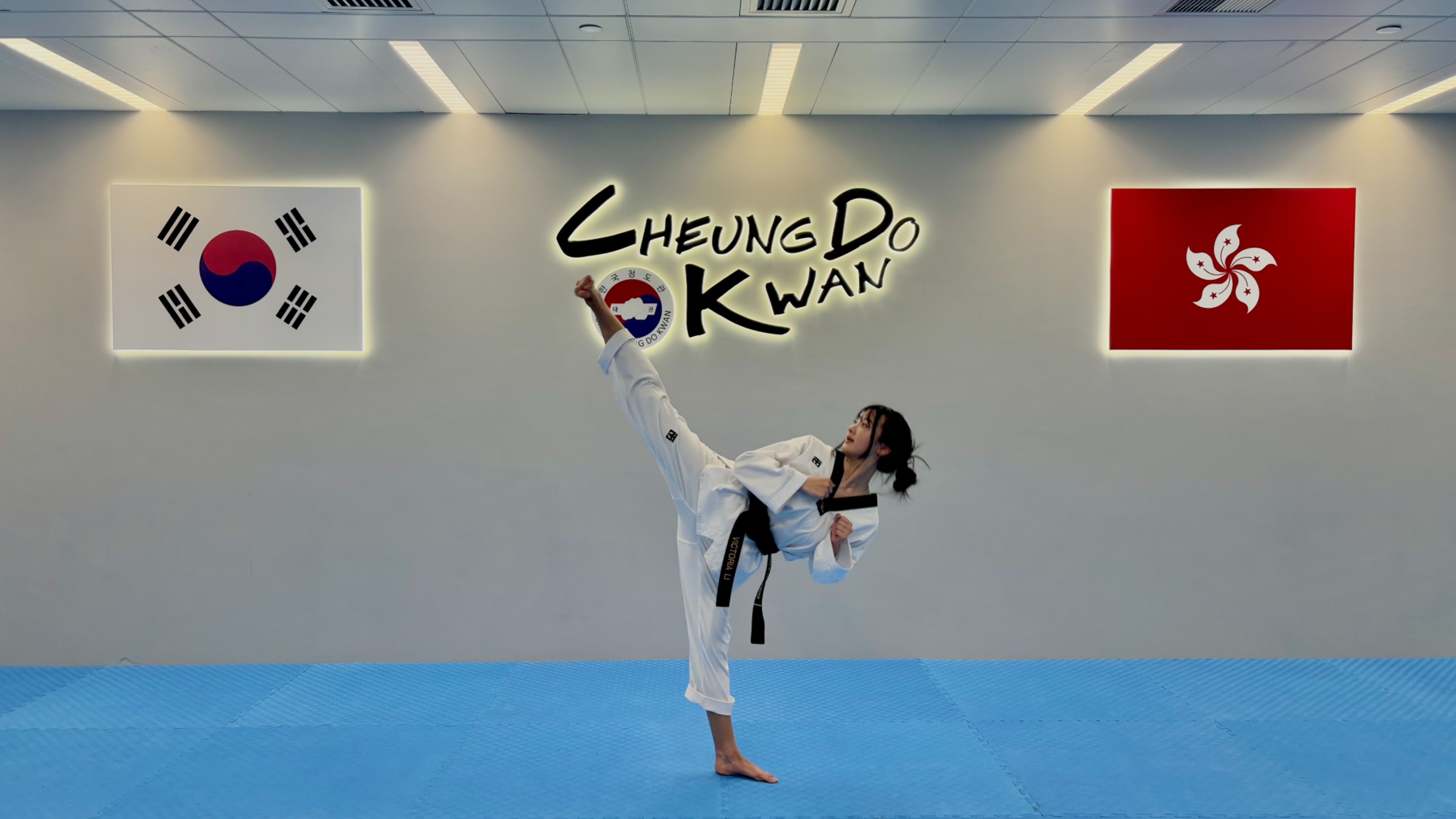 Young woman, solitary on a blue mat, wearing Taekwondo uniform kicks into the air.