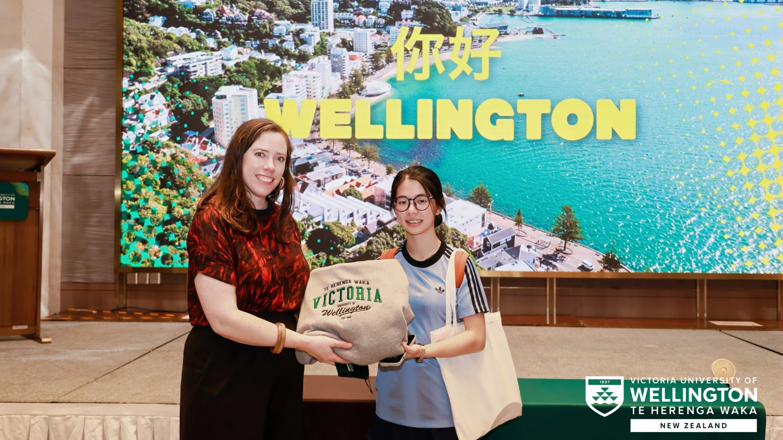 White woman with long dark hair and red top handing a hoodie with 'Victoria' to a Chinese girl with glasses and a grey top