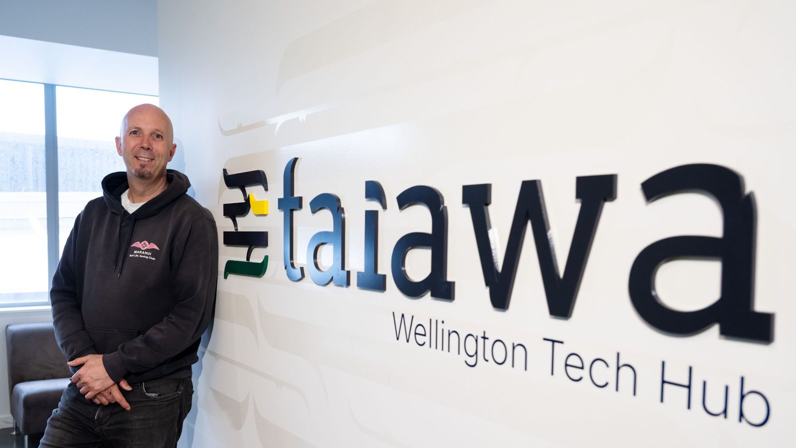 Bald man in hoodie, posing against a wall with a sign that says 'Taiawa'