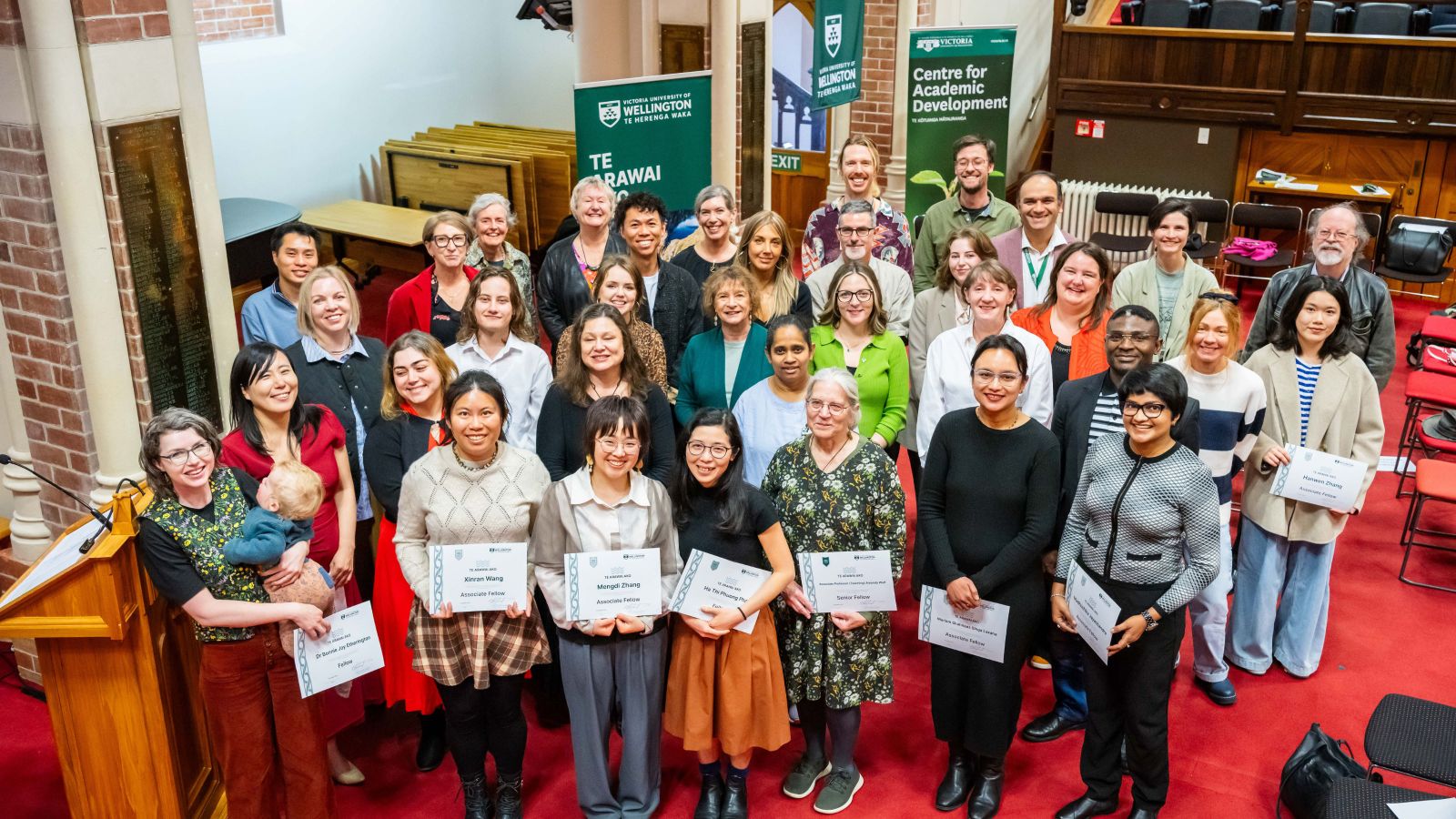 A large group of people stand in Council Chamber, holding their certificates.
