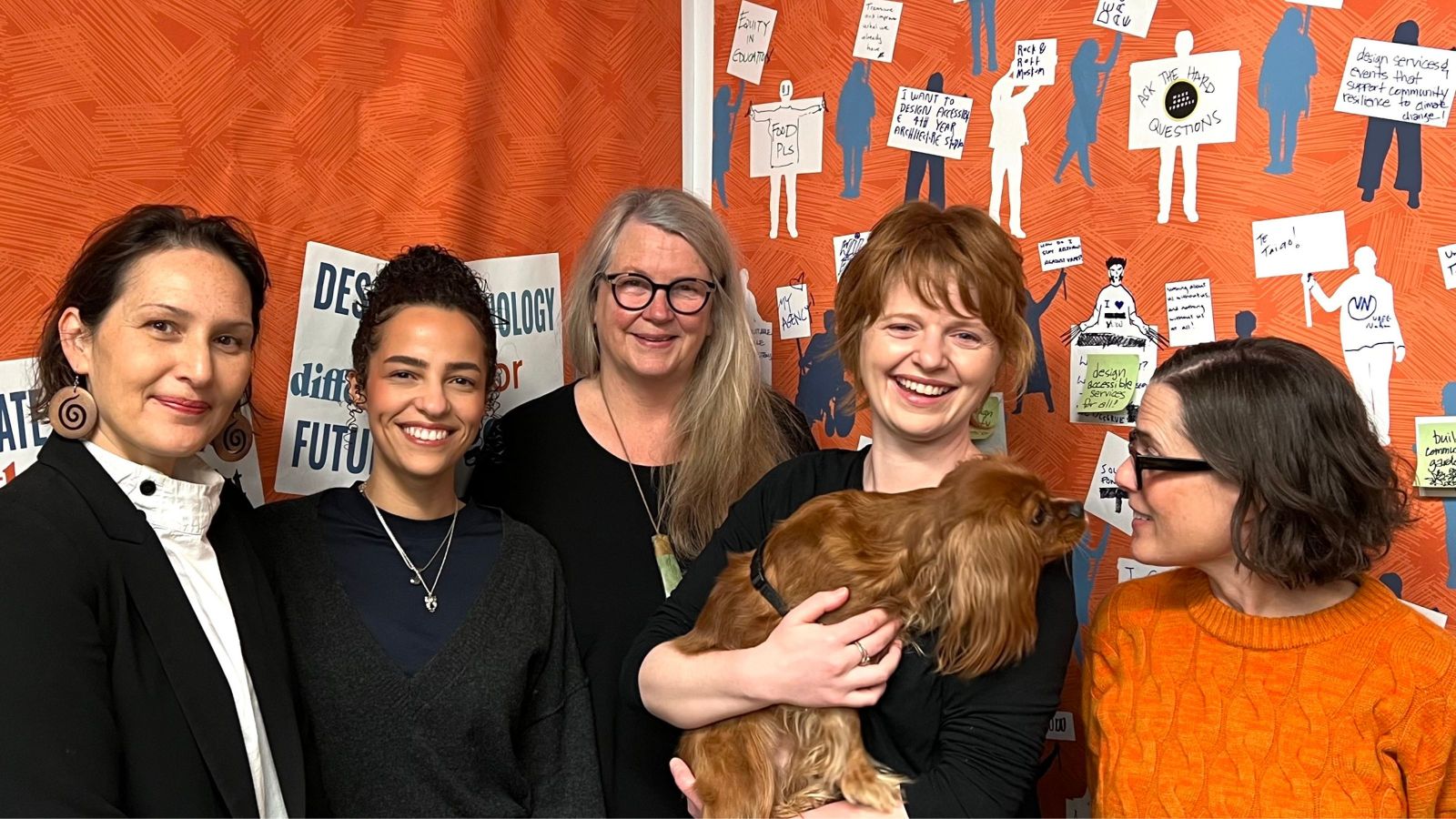 Four people and a dog in front of an orange background with statements about design written all over it