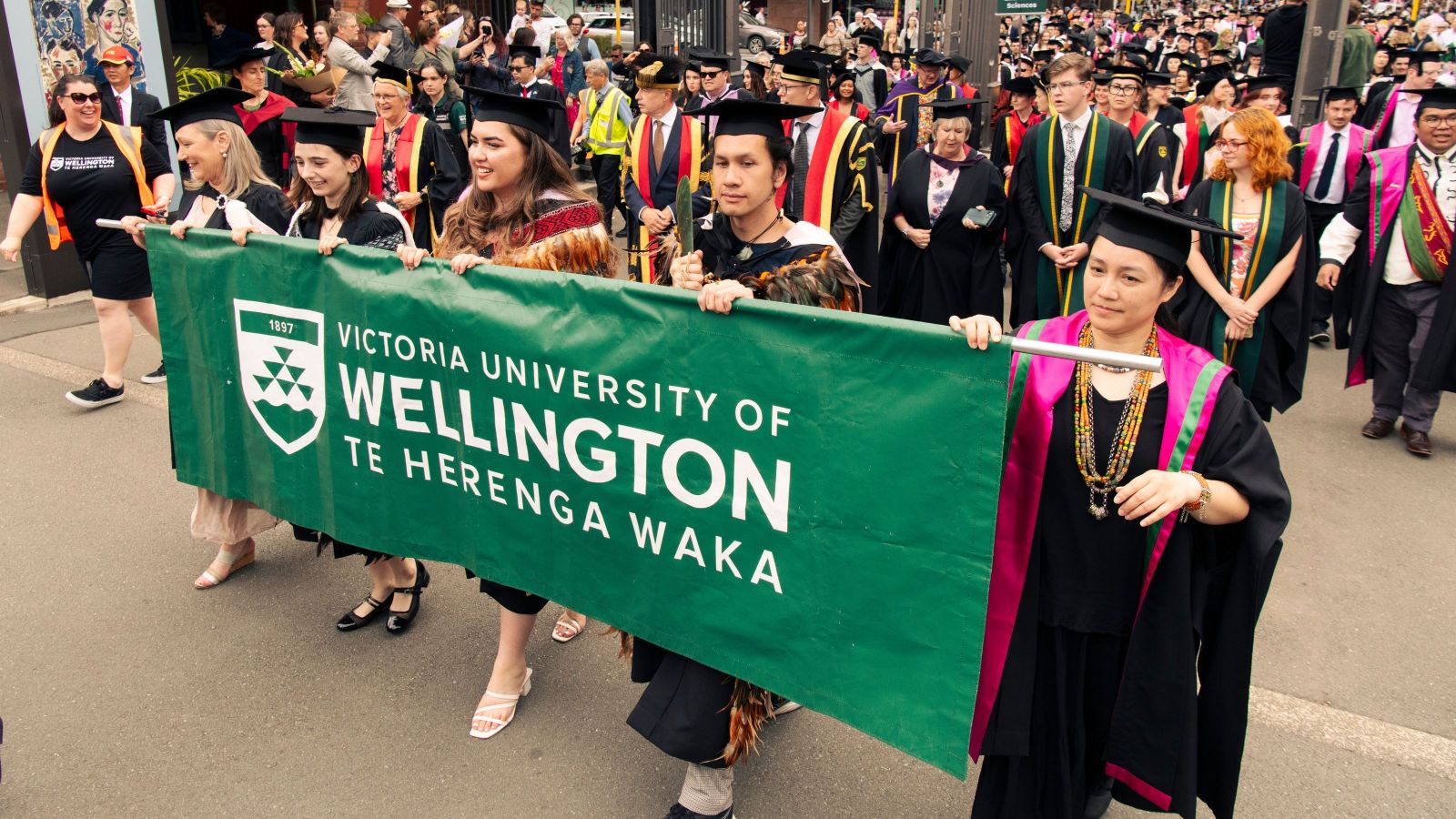 four people in graduation gear holding a wide dark green banner with university name on