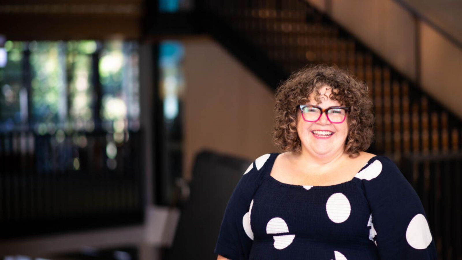 Woman with curly hair and pink and purple glasses, wearing a black and white large polka dot top