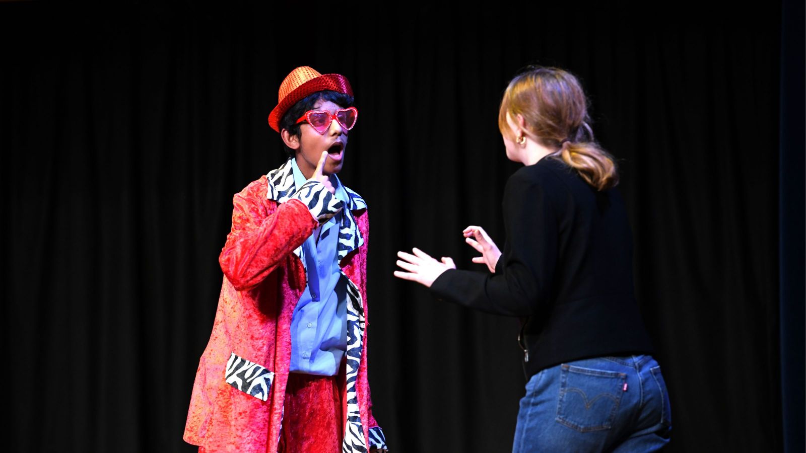 two people on a dark stage with spotlight, one wearing red crushed velvet standing with his finger to his mouth, the other in black with blue jeans