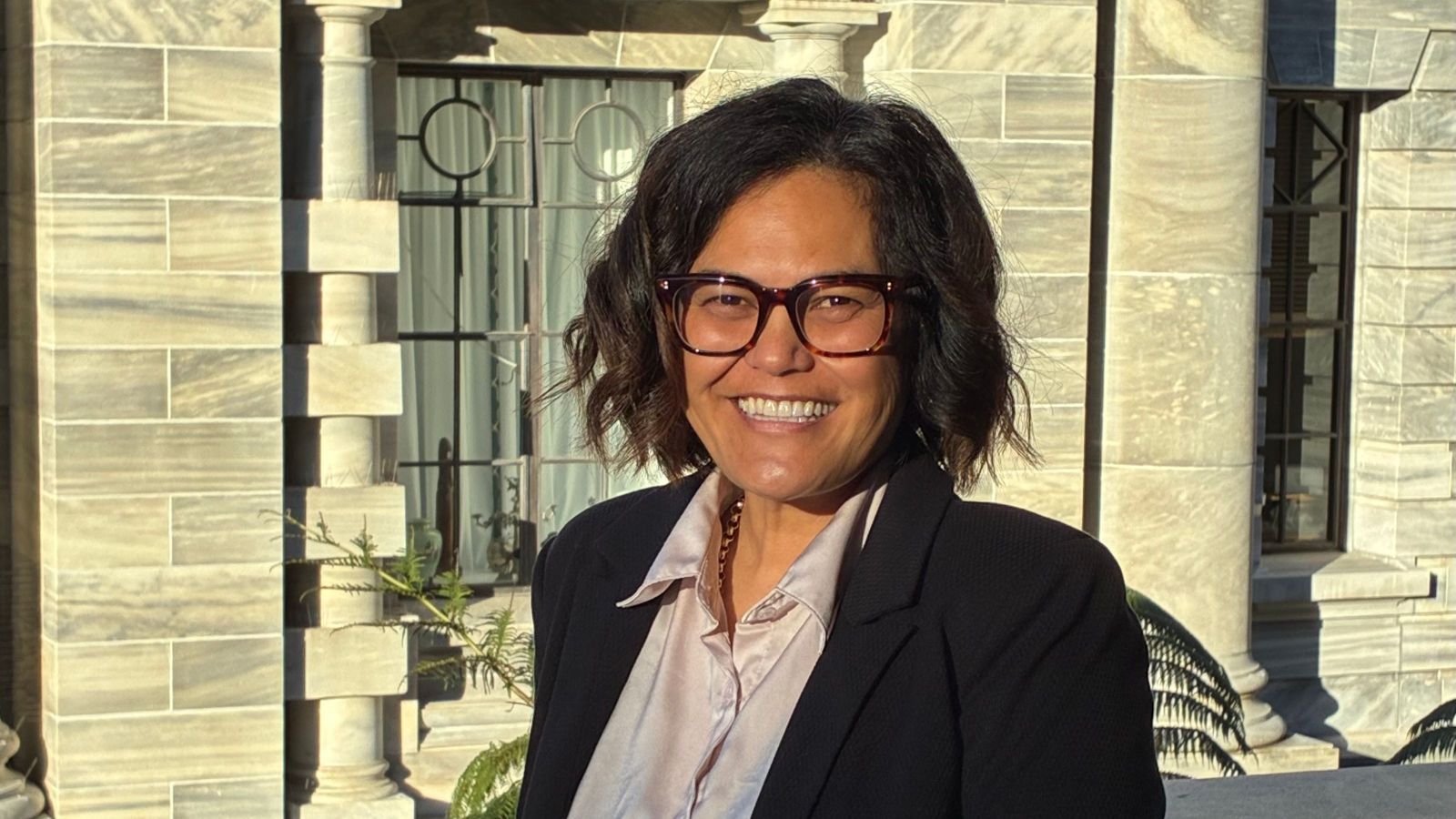 Woman with dark-framed eyeglasses, curly bobbed hair against building with grey stone architecture