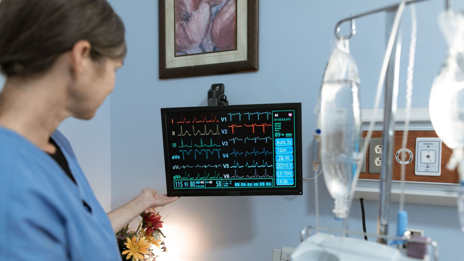 A nurse in blue scrubs looking at a medical monitor next to a hospital bed 
