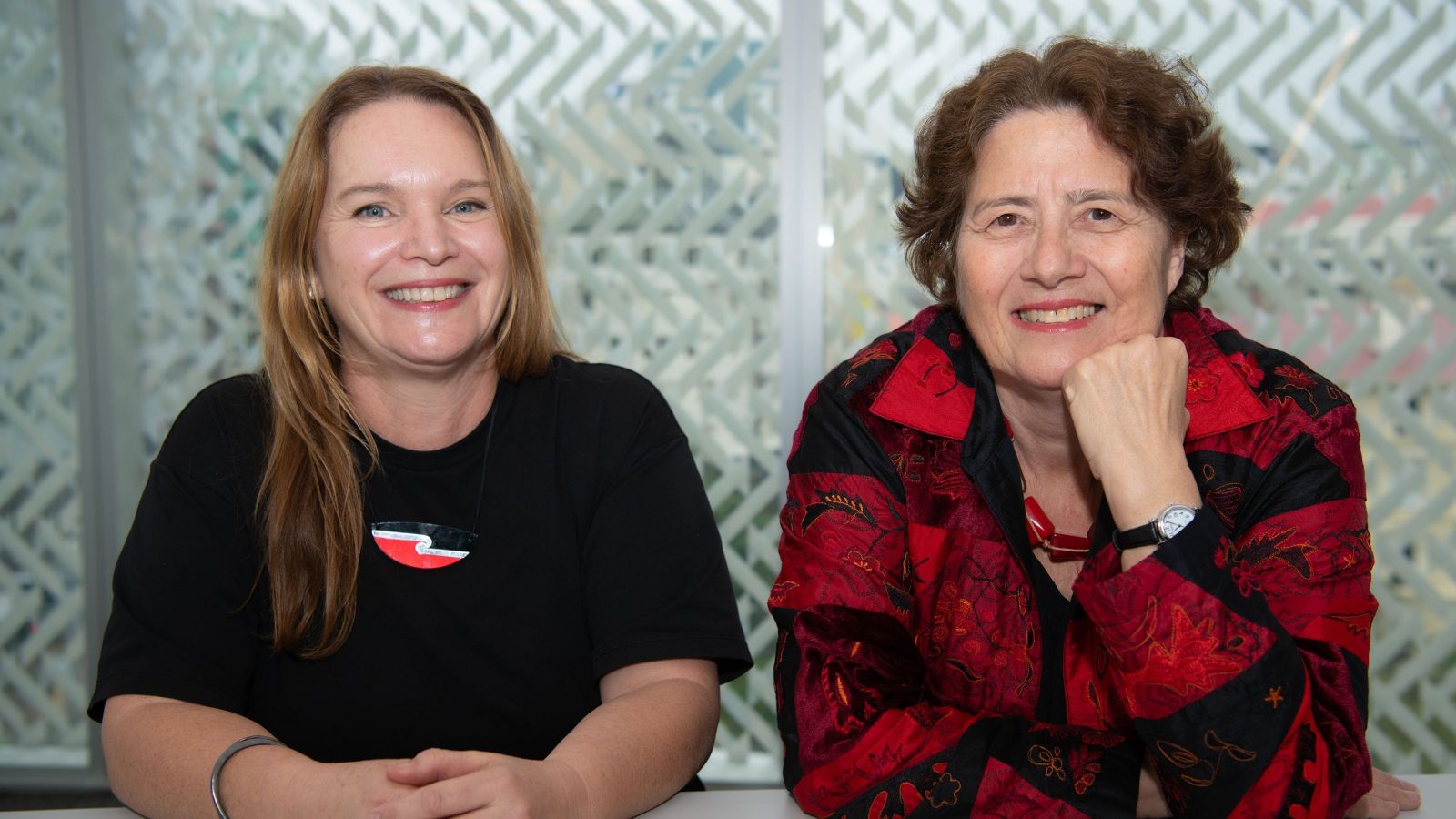two women sitting alongside each other, one with long blond hair and blue eyes, one with curly hair and dark eyes