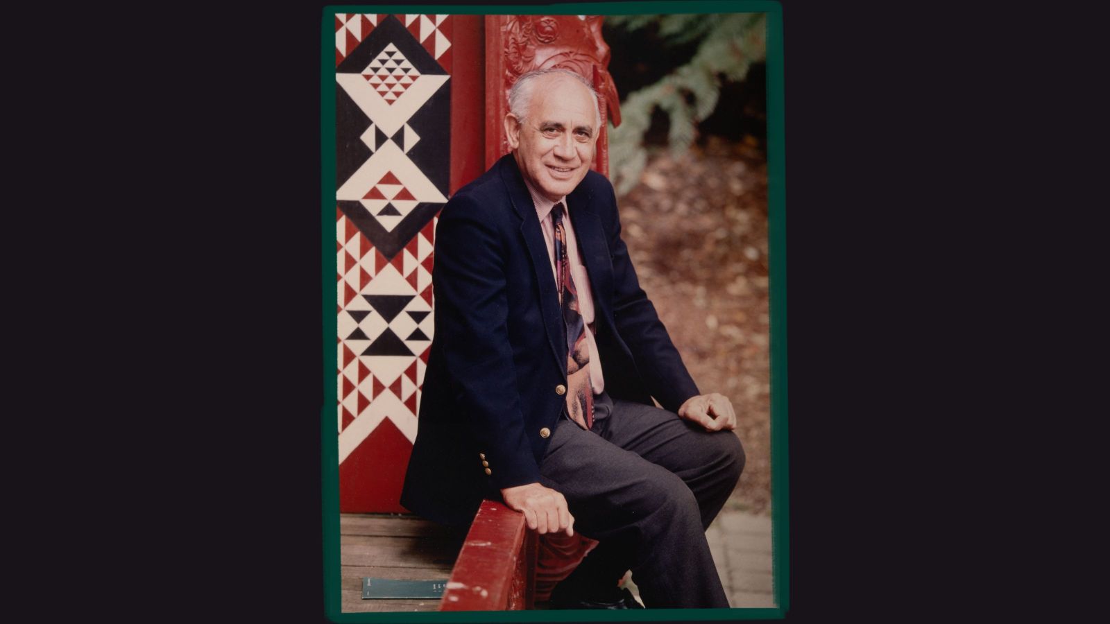 Man sitting on the verandah of a marae with red carvings in the background