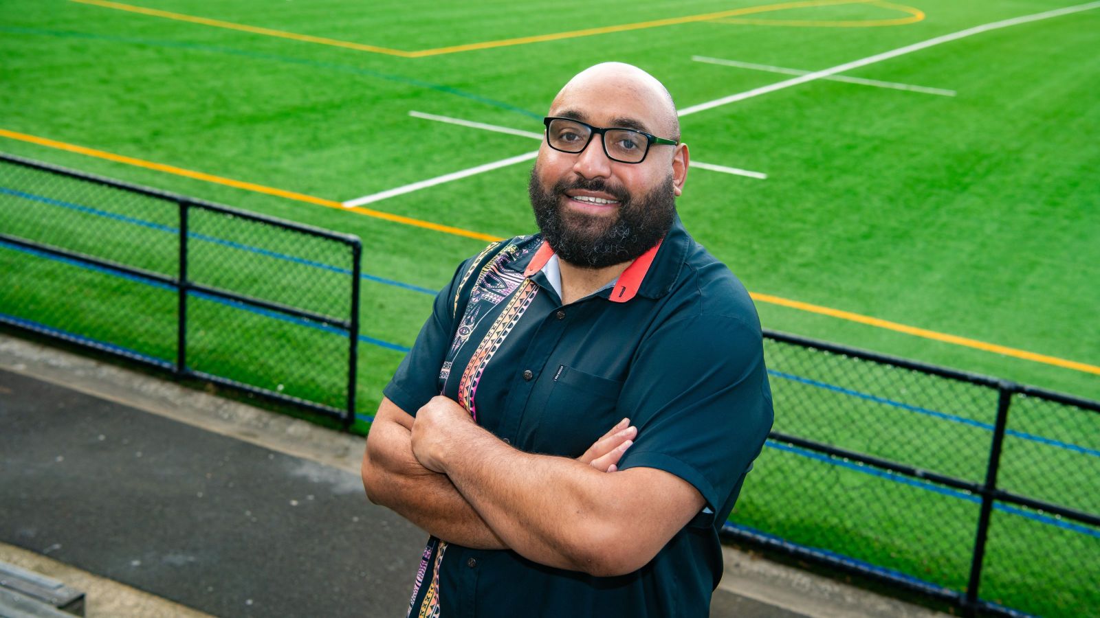 image of man in a samoan style top leaning against a fence in front of a bright green turf