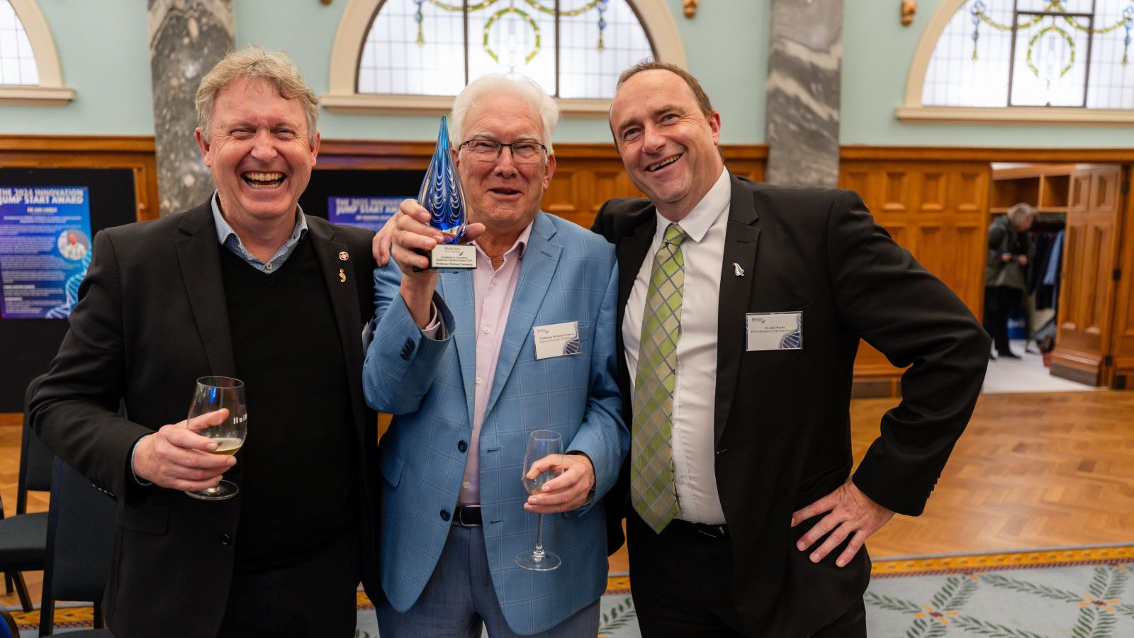 Three men looking delighted, two with dark suits, the centre one with a light blue check suit