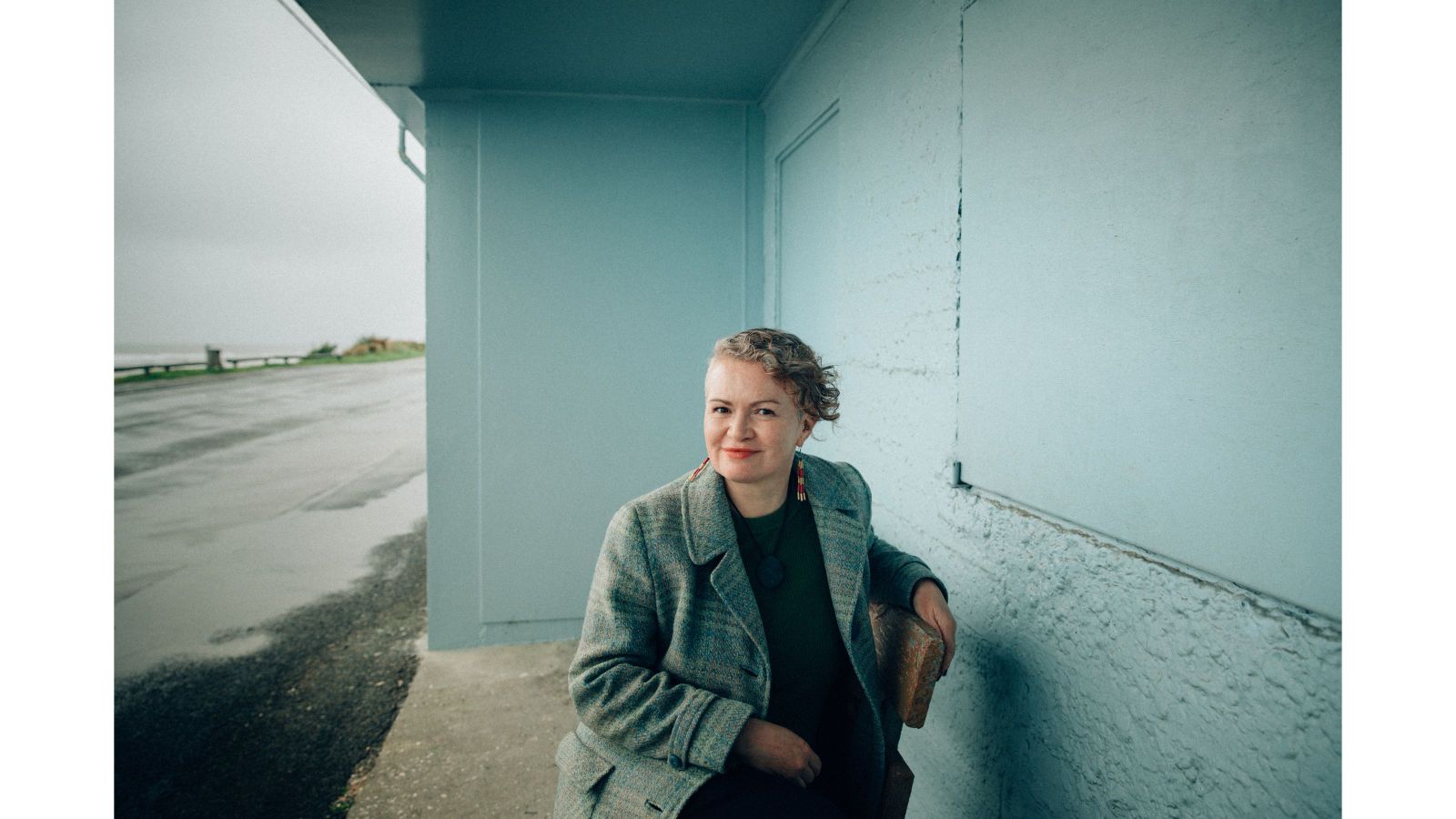 woman with funky short hair in pale blue bus stop with ocean in background