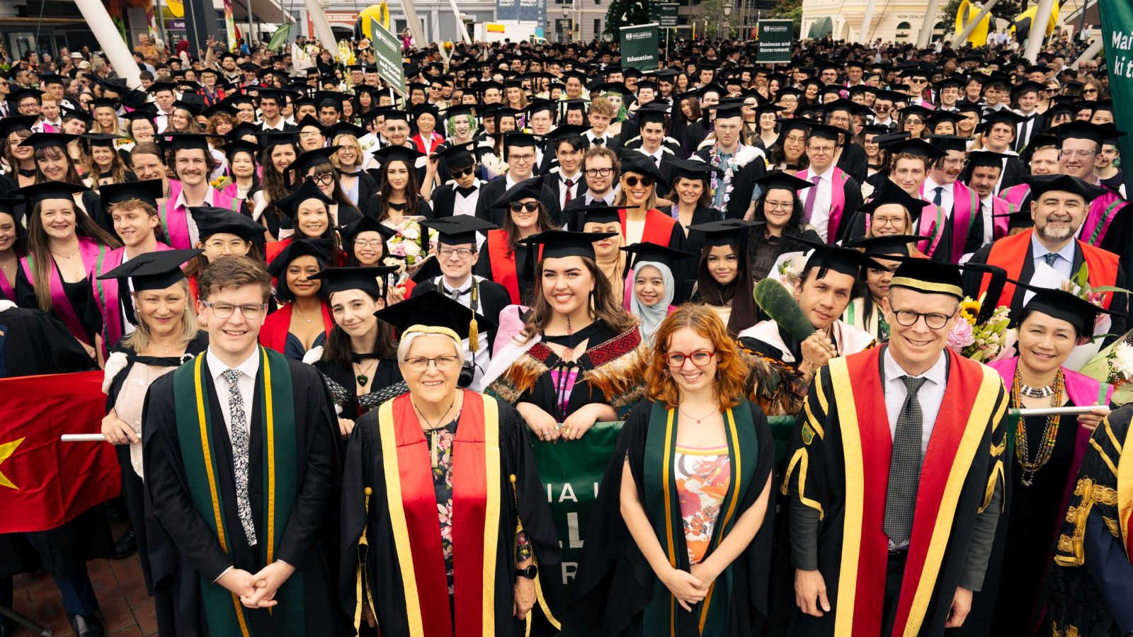 Hundreds of students in academic gowns, hoods, and caps smile for the camera
