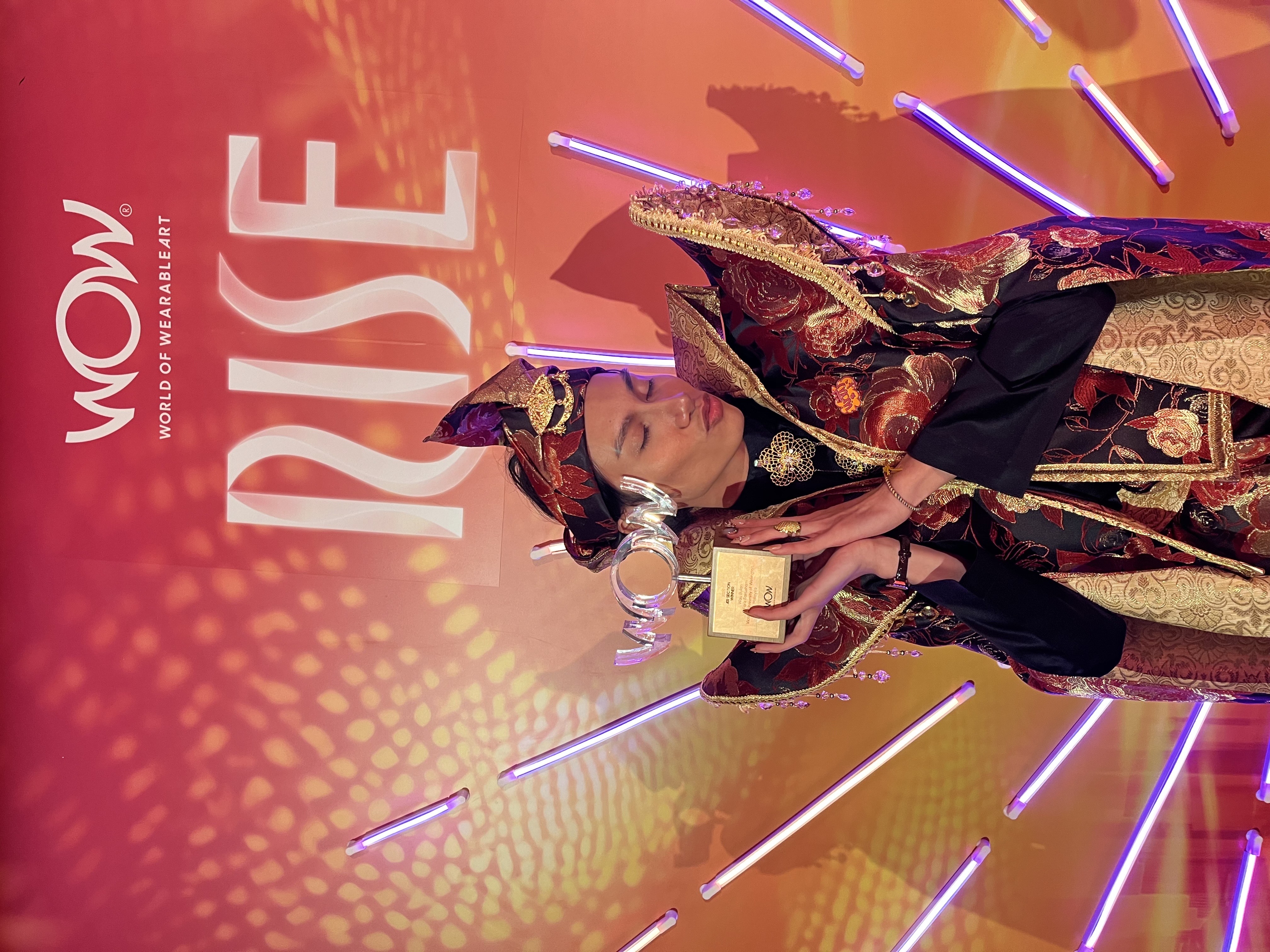 Faris wears handmade garment, holding award.