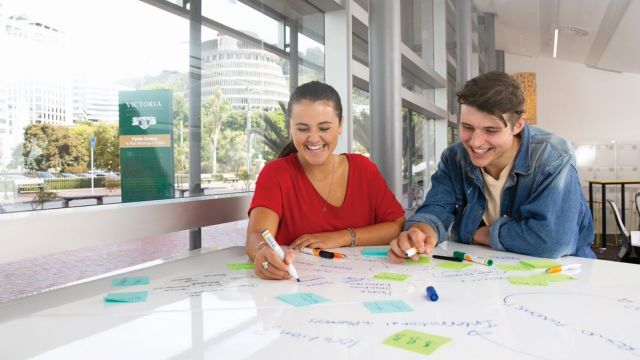 A male and female student, draw on white board with markers at a table.