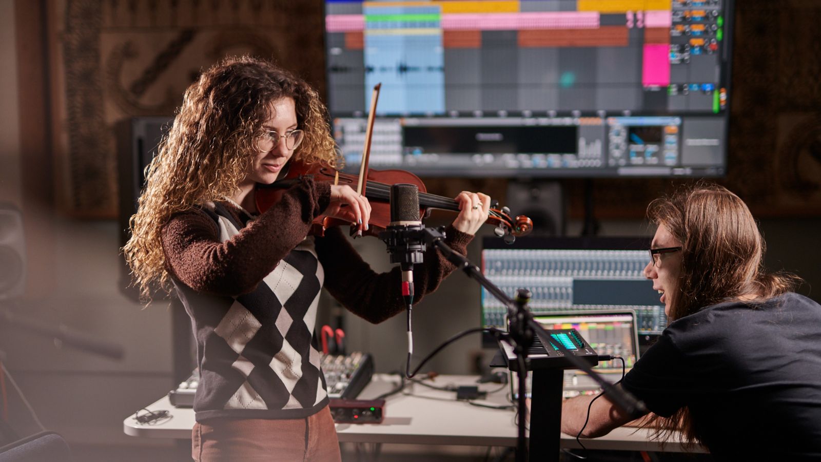 Two students in a recording studio, one is playing the violin, and the other is sat at the sound board.