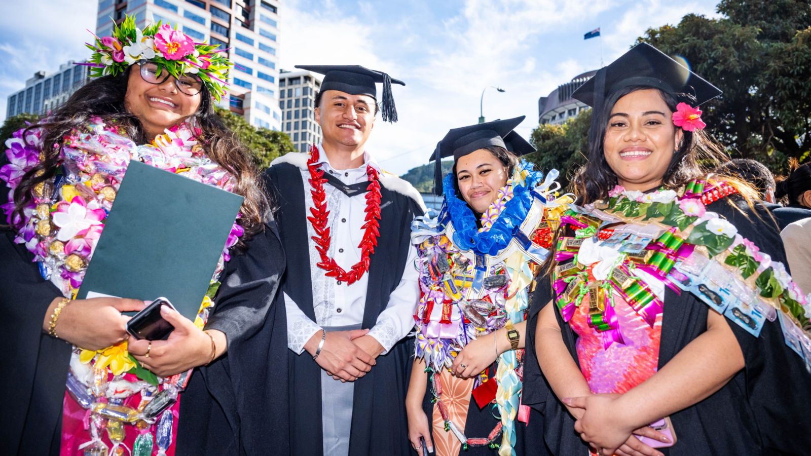 Four graduates in caps and gowns with leis, posing outdoors.