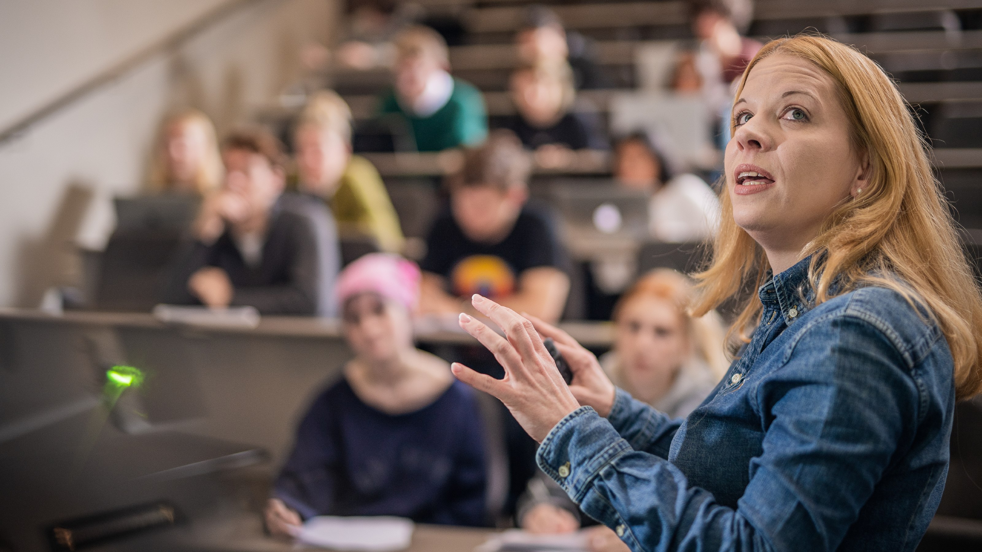 A female lecturer half-turns away from the audience to view an unseen screen behind her.
