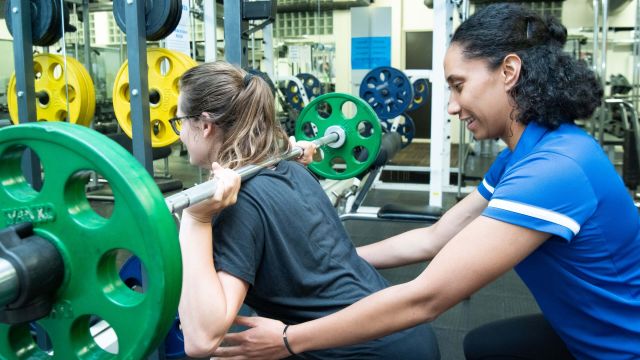 A woman is lifting a heavy weight while being monitored by a personal trainer. 