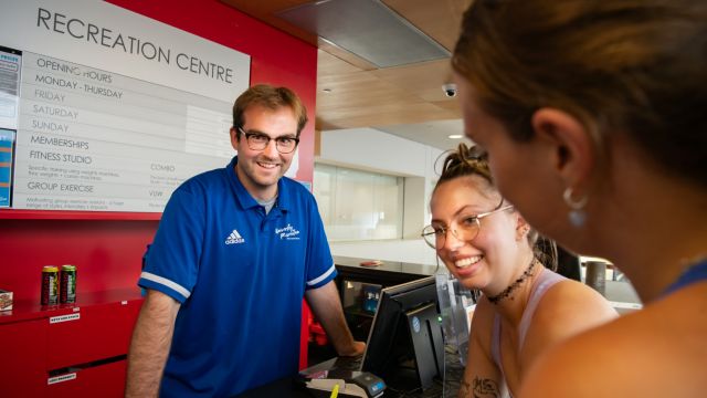 A smiling staff member in a blue polo shirt assists two people at the reception desk of a recreation centre, with a sign displaying opening hours in the background.