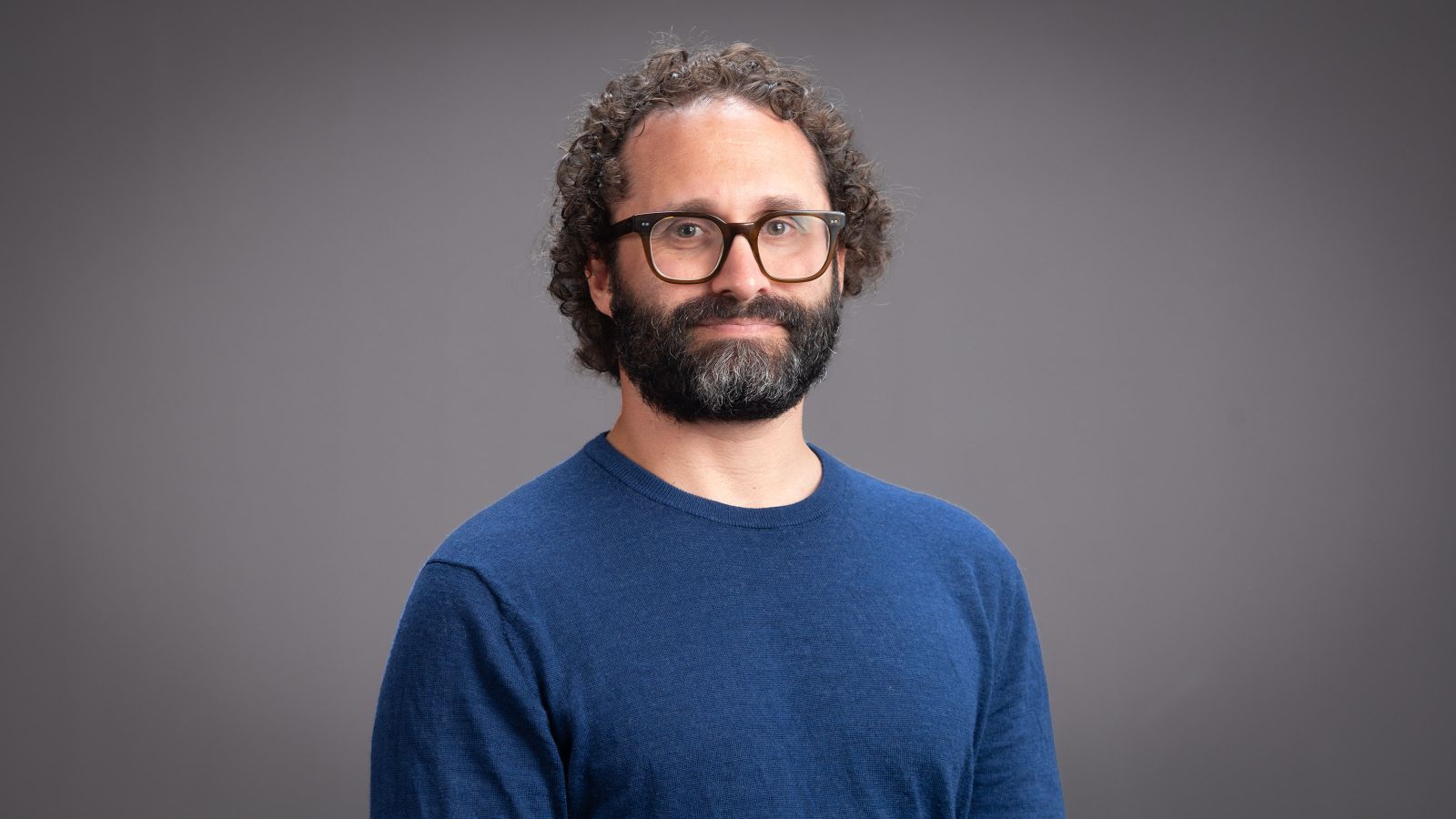 A man with short curly black hair, a black beard and mustache wearing glasses and a blue top smiles at the camera