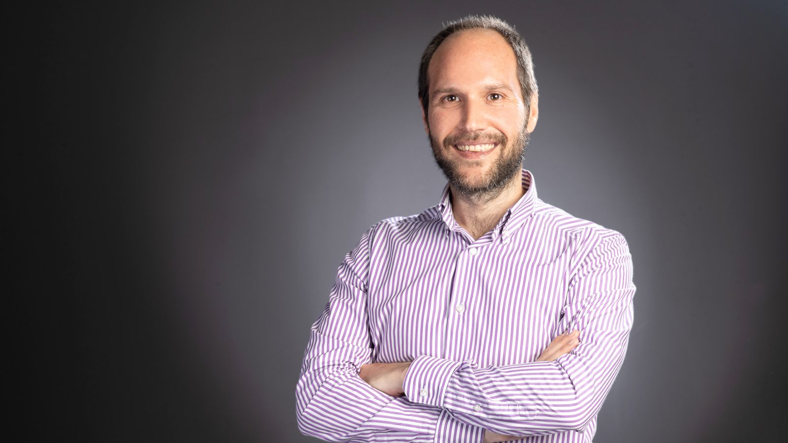 A man with short grey hair and a beard wearing a pink shirt stands with his arms crossed and smiles at the camera