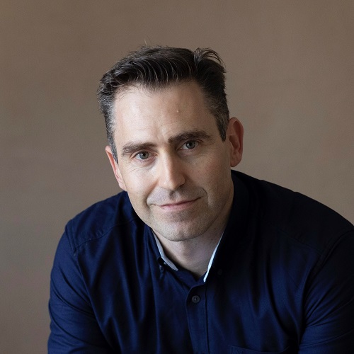 A man with dark hair wearing a navy blue shirt smiles at the camera