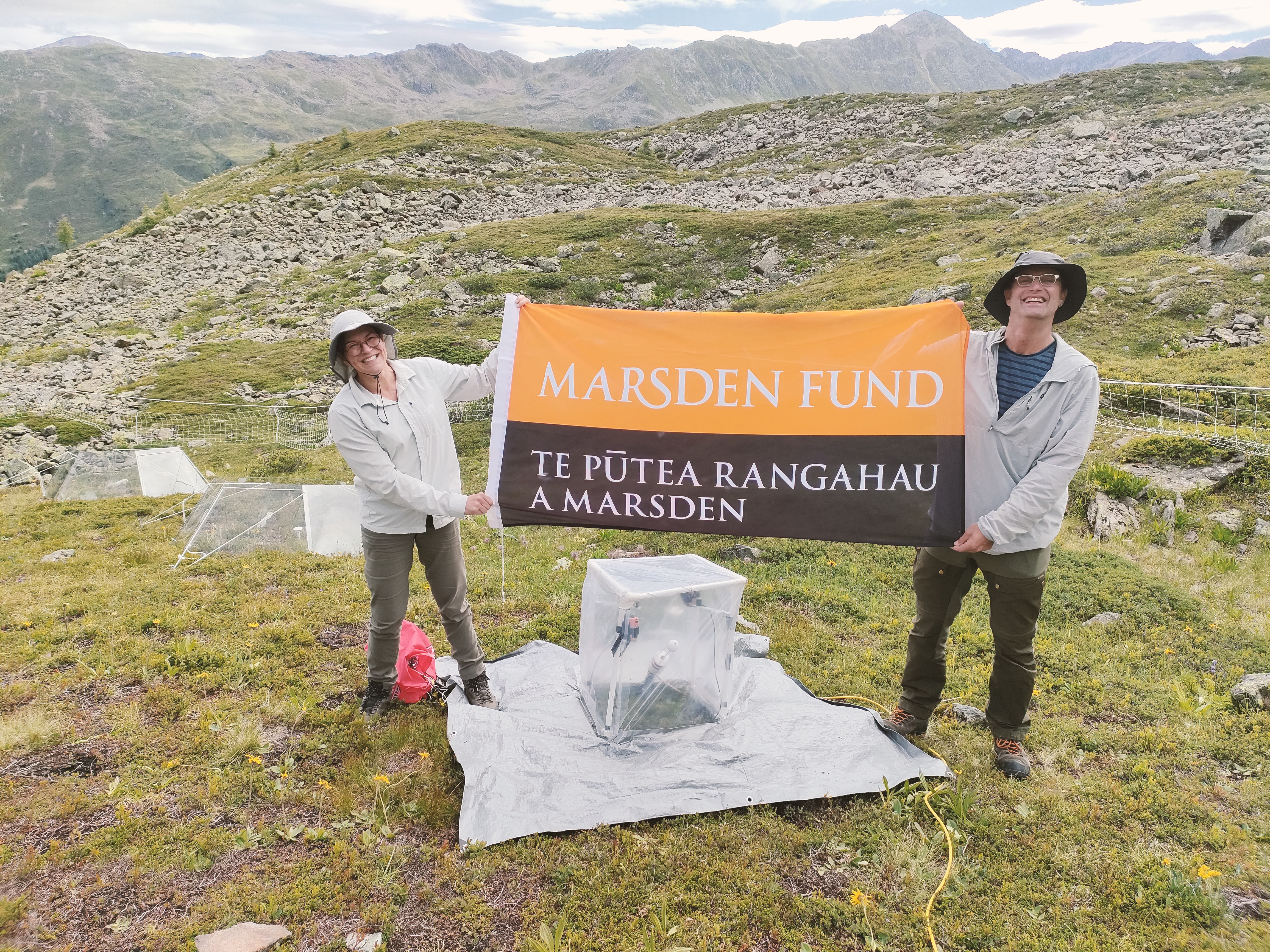 Two people standing in alpine grassland holding an orange and black banner reading “Marsden Fund – Te Pūtea Rangahau a Marsden,” with rocky hills and mountains in the background and research equipment set up on the ground in front of them.