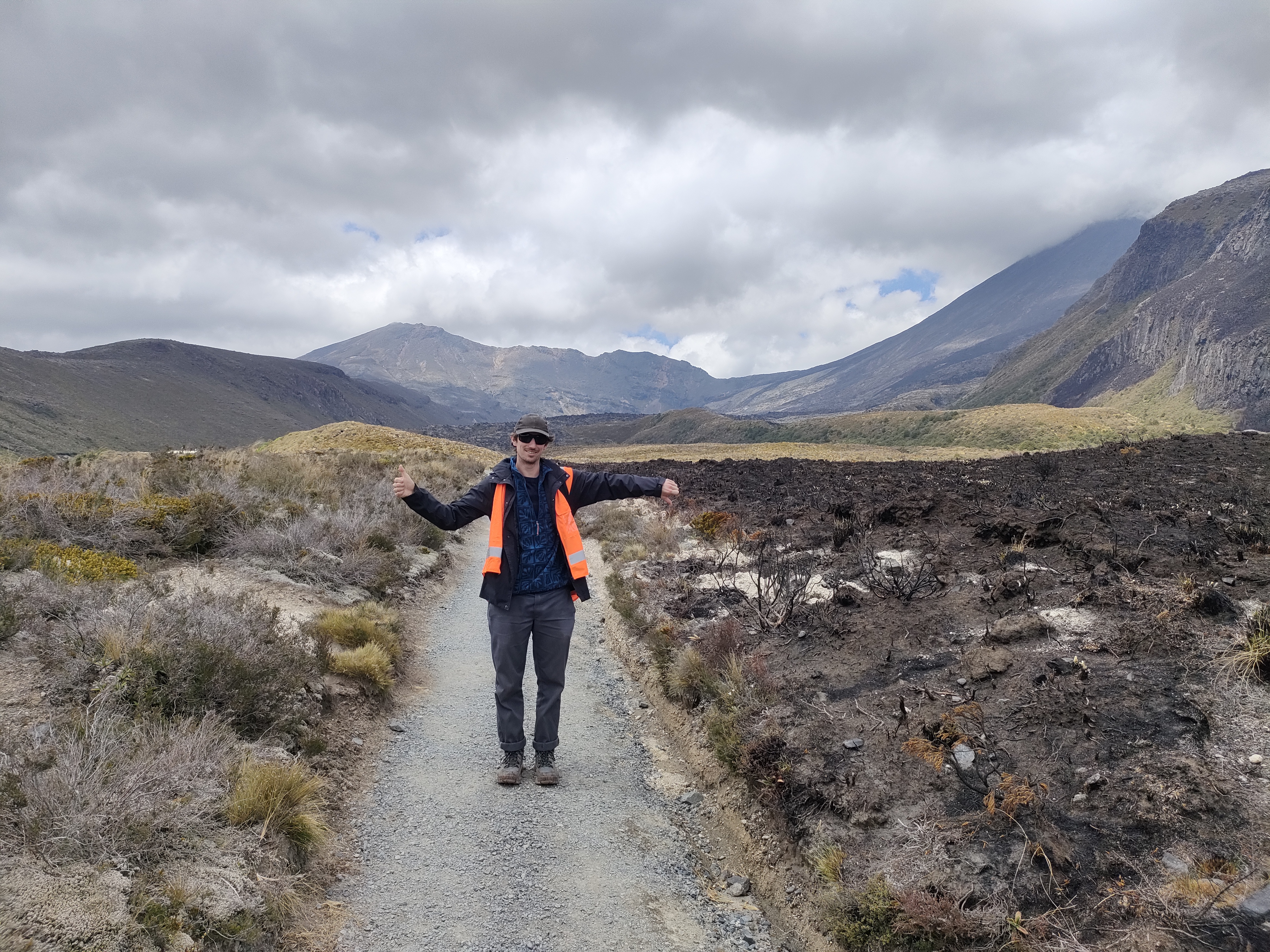Person standing on a gravel track with arms outstretched, giving a thumbs up on the left side beside unburnt alpine vegetation and a thumbs down on the right side beside dark, recently burnt ground, with mountains under a cloudy sky in the background.
