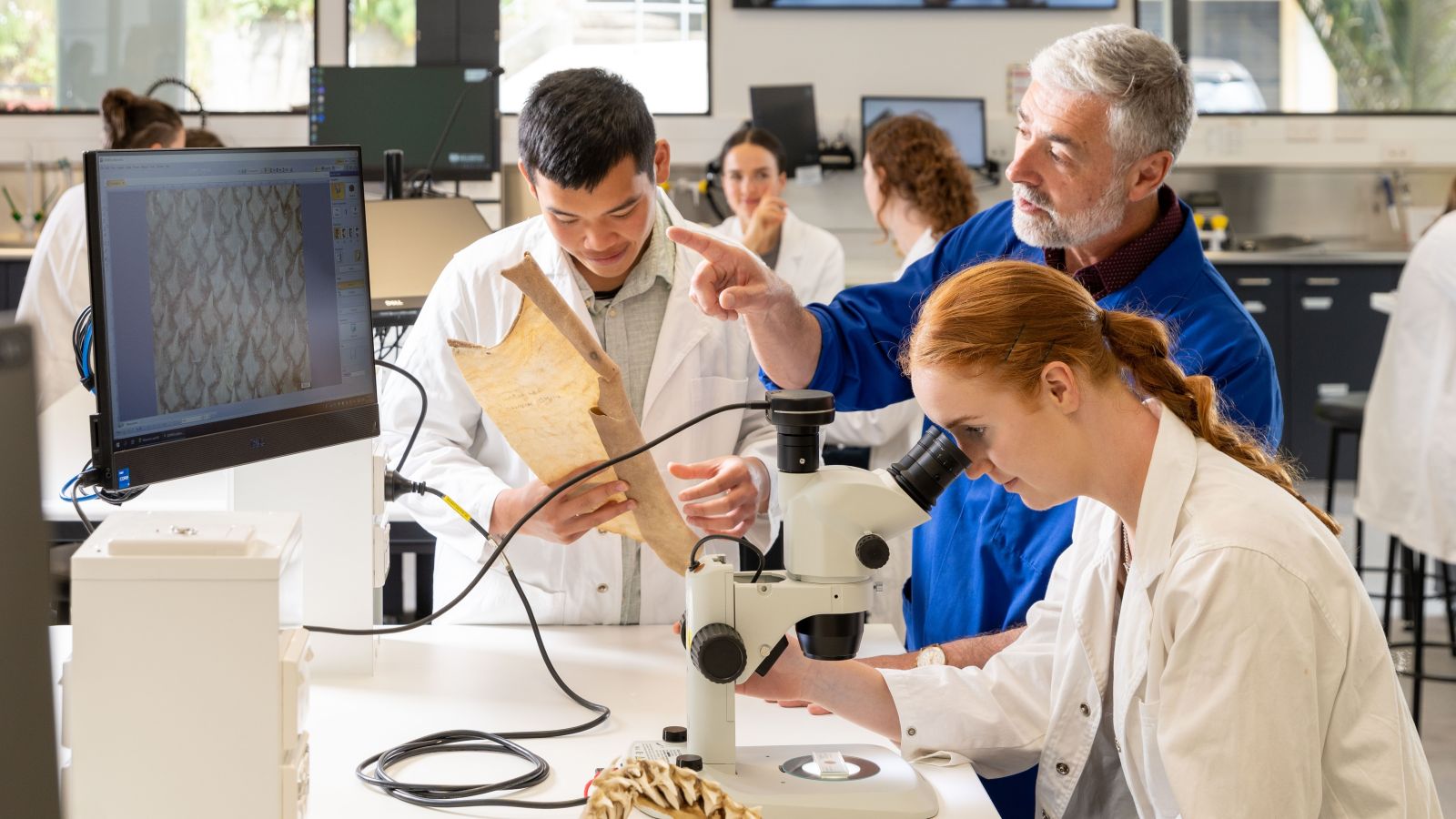 Biomedical Sciences students use a microscope and digital imaging equipment to examine biological specimens in a teaching laboratory. One student observes a specimen through a microscope while another student and an instructor examine a sample and discuss observations. A computer monitor displays a magnified image of the specimen being studied.